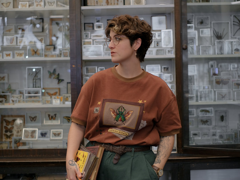 A rusty, vintage orange cotton T-shirt featuring an embroidered stag-horn beetle design framed with vintage-style details, handwritten notes, celestial accents, and latin nomenclature.