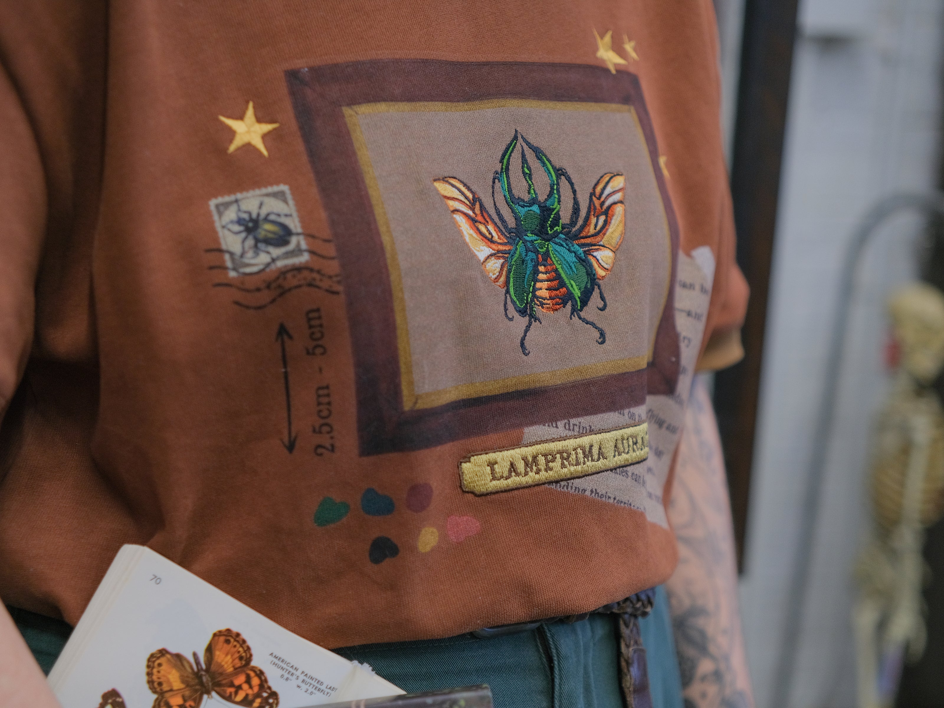 A rusty, vintage orange cotton T-shirt featuring an embroidered stag-horn beetle design framed with vintage-style details, handwritten notes, celestial accents, and latin nomenclature.