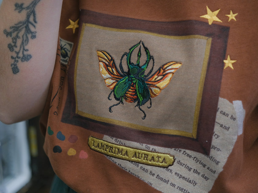 A rusty, vintage orange cotton T-shirt featuring an embroidered stag-horn beetle design framed with vintage-style details, handwritten notes, celestial accents, and latin nomenclature.