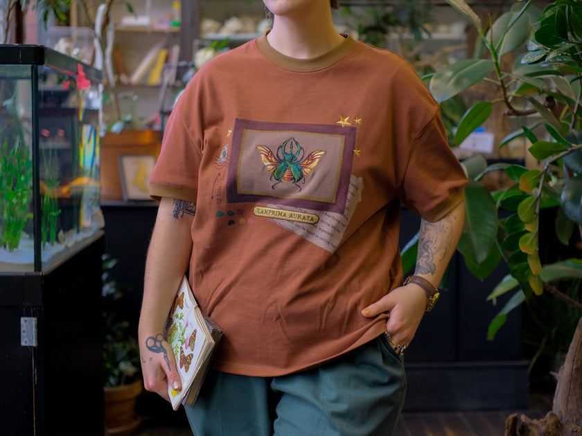 A rusty, vintage orange cotton T-shirt featuring an embroidered stag-horn beetle design framed with vintage-style details, handwritten notes, celestial accents, and latin nomenclature.