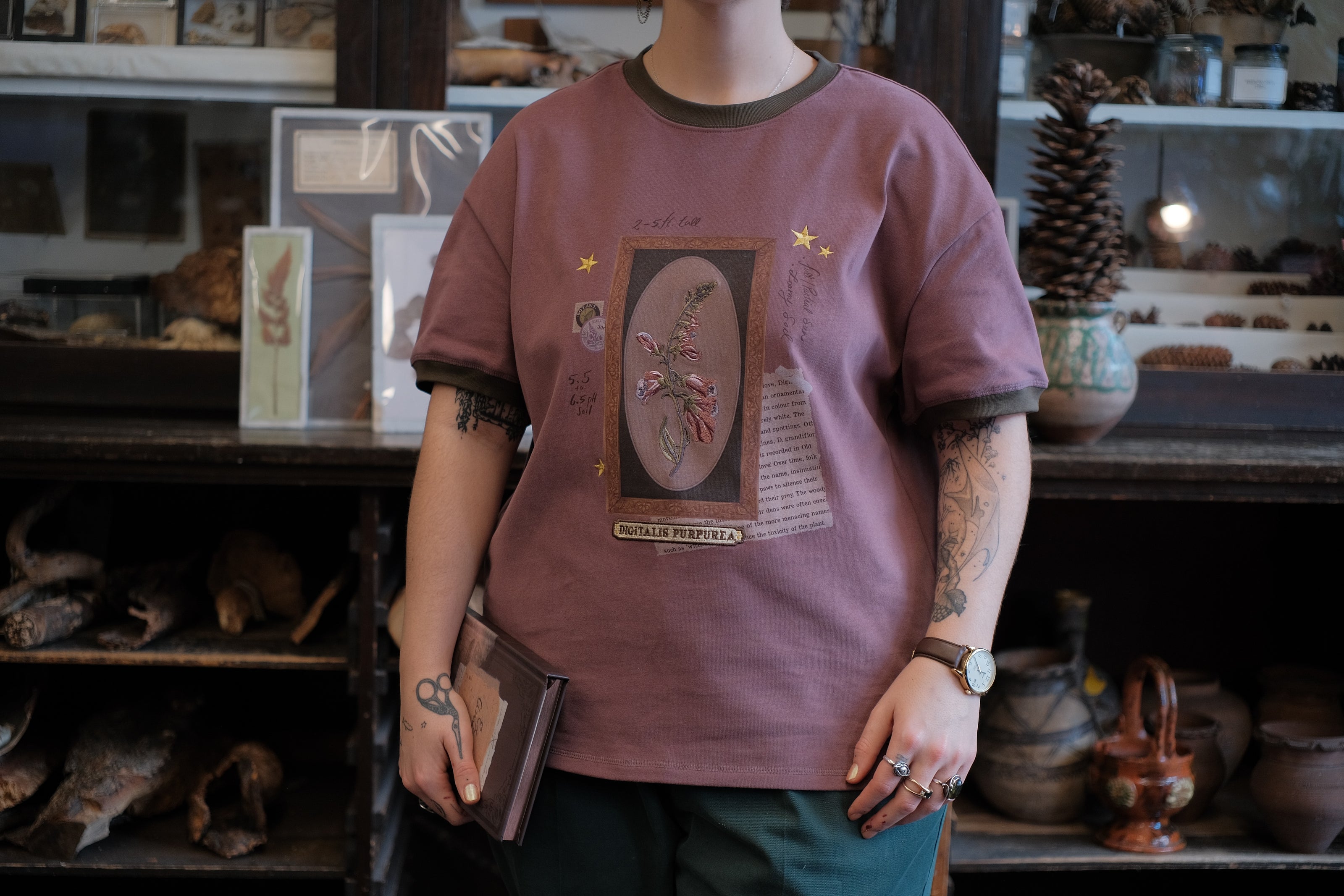 Mauve-coloured cotton T-shirt featuring a detailed botanical illustration of a Digitalis Purpurea (foxglove) plant framed within an ornate oval design. The shirt is embellished with handwritten notes, stars, and vintage-style accents, creating a natural and artistic aesthetic.