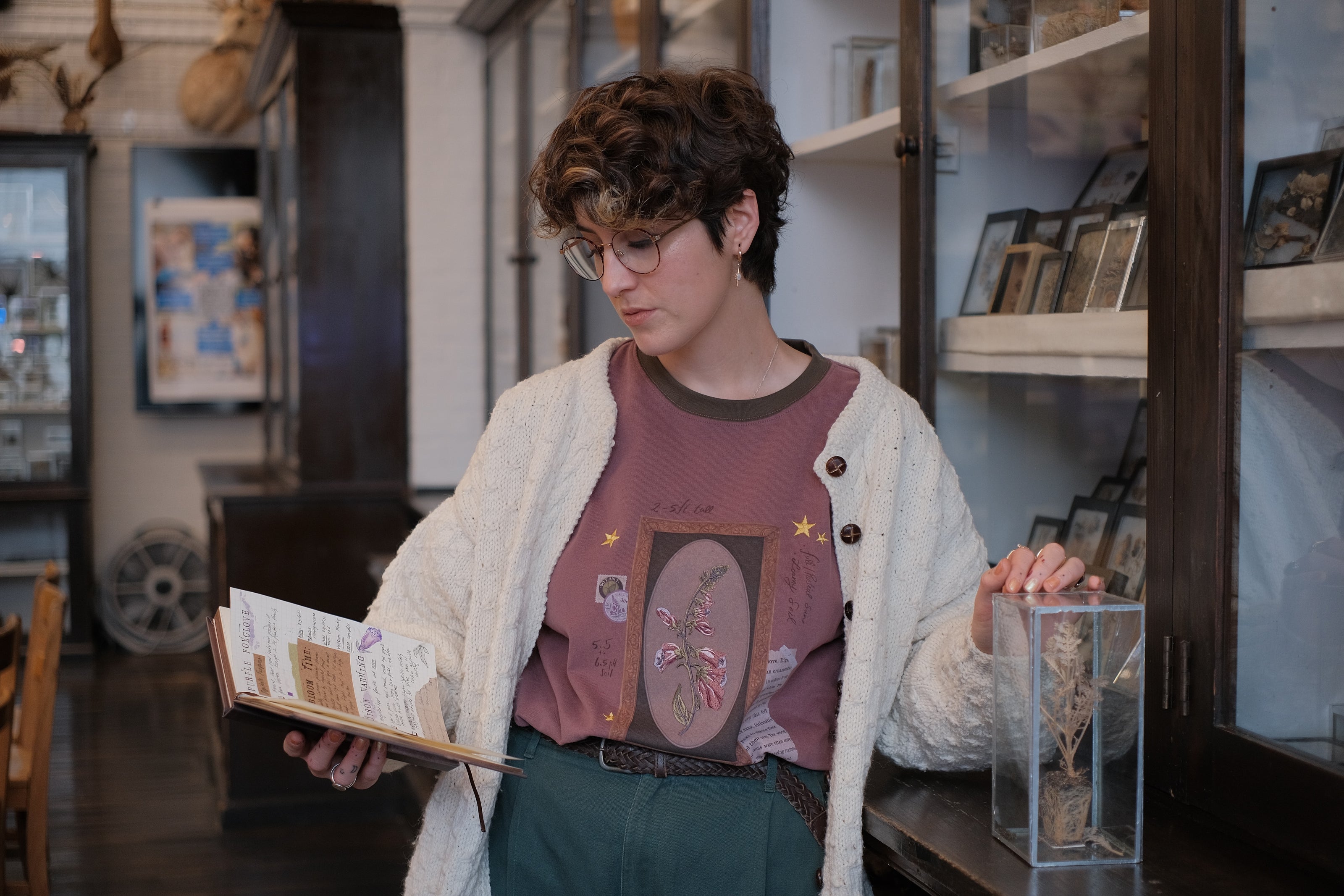 Mauve-coloured cotton T-shirt featuring a detailed botanical illustration of a Digitalis Purpurea (foxglove) plant framed within an ornate oval design. The shirt is embellished with handwritten notes, stars, and vintage-style accents, creating a natural and artistic aesthetic.