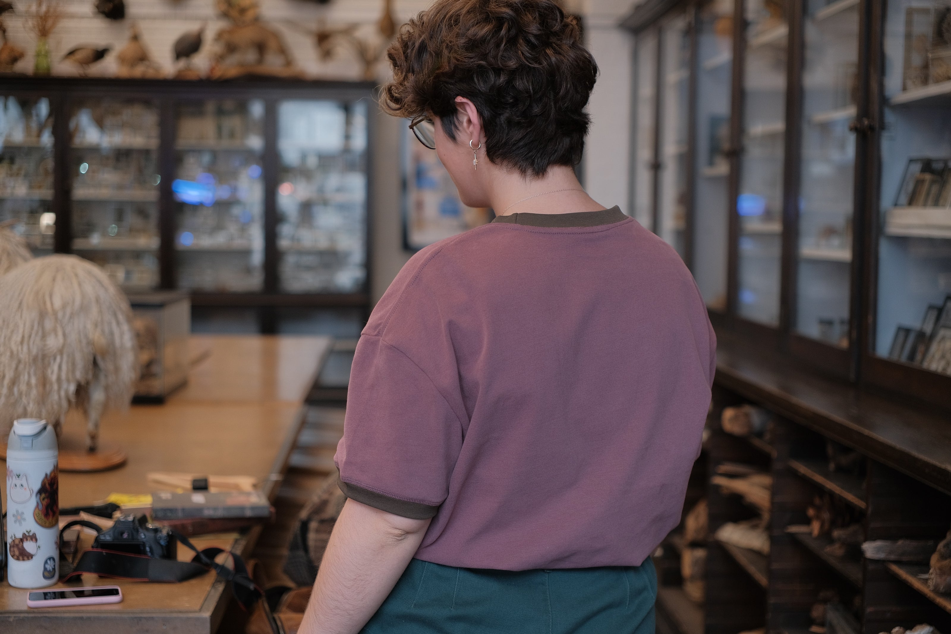 Mauve-coloured cotton T-shirt featuring a detailed botanical illustration of a Digitalis Purpurea (foxglove) plant framed within an ornate oval design. The shirt is embellished with handwritten notes, stars, and vintage-style accents, creating a natural and artistic aesthetic.