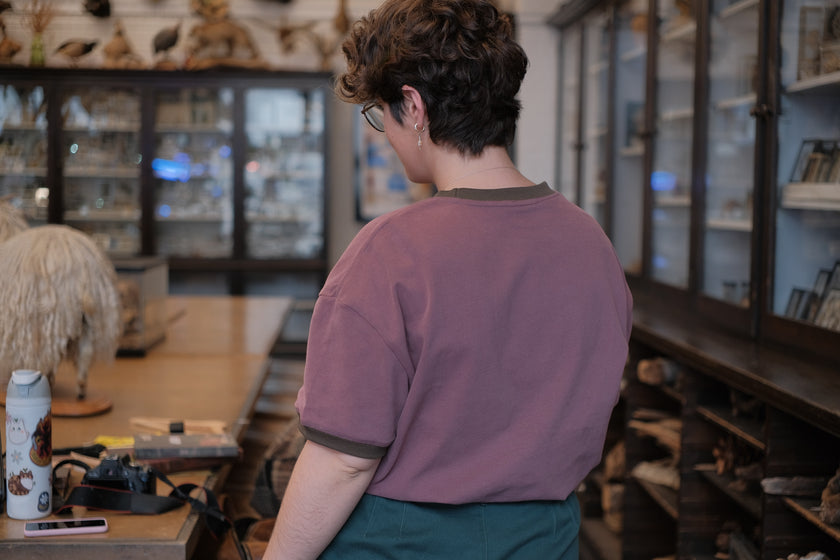 Mauve-coloured cotton T-shirt featuring a detailed botanical illustration of a Digitalis Purpurea (foxglove) plant framed within an ornate oval design. The shirt is embellished with handwritten notes, stars, and vintage-style accents, creating a natural and artistic aesthetic.