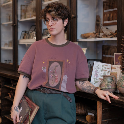 Mauve-coloured cotton T-shirt featuring a detailed botanical illustration of a Digitalis Purpurea (foxglove) plant framed within an ornate oval design. The shirt is embellished with handwritten notes, stars, and vintage-style accents, creating a natural and artistic aesthetic.