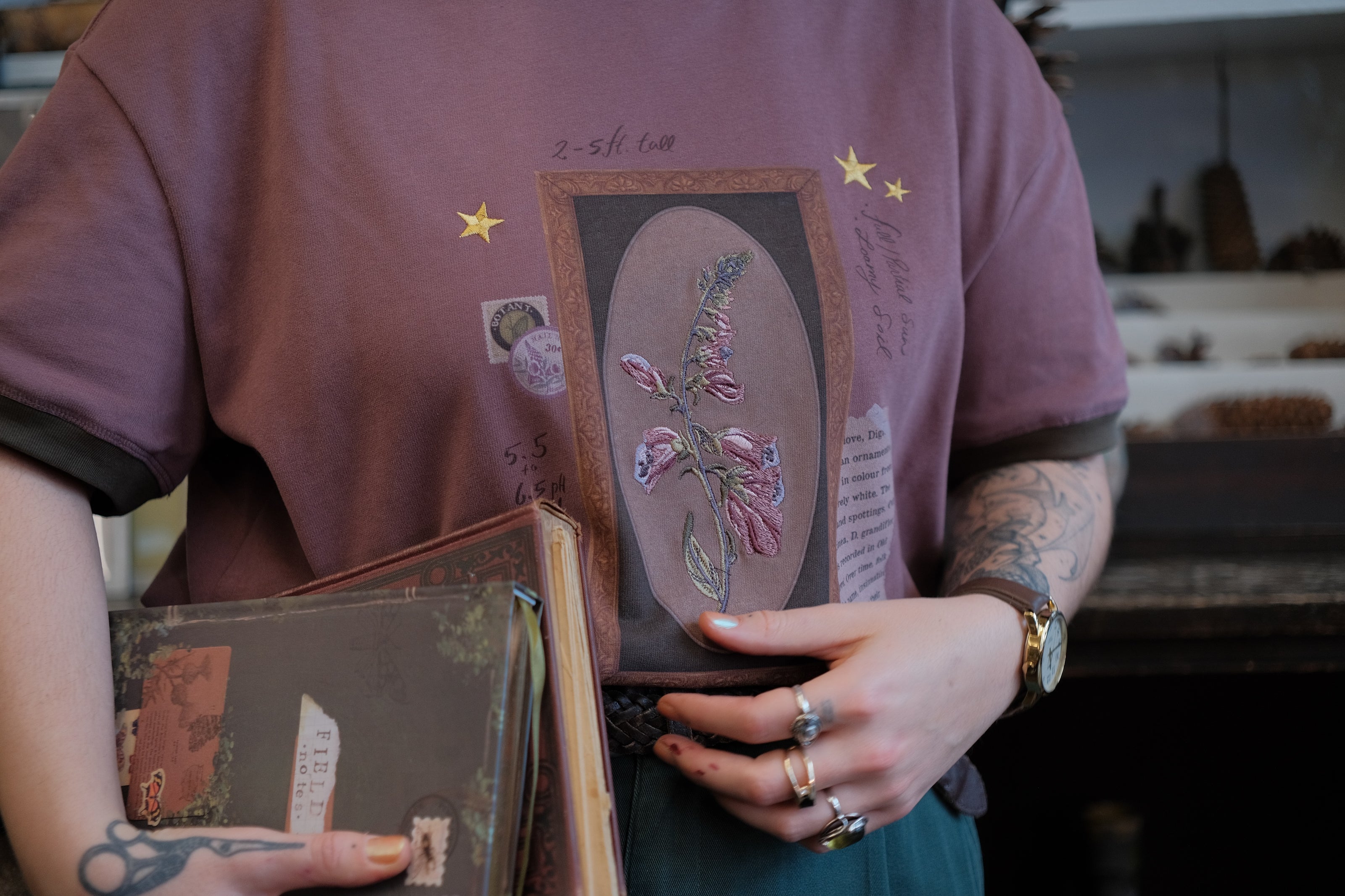 Mauve-coloured cotton T-shirt featuring a detailed botanical illustration of a Digitalis Purpurea (foxglove) plant framed within an ornate oval design. The shirt is embellished with handwritten notes, stars, and vintage-style accents, creating a natural and artistic aesthetic.