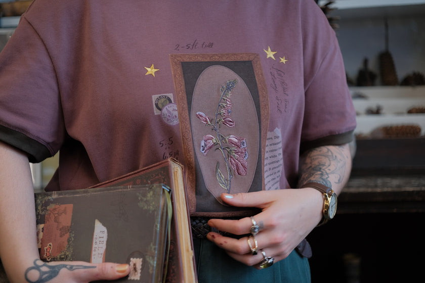 Mauve-coloured cotton T-shirt featuring a detailed botanical illustration of a Digitalis Purpurea (foxglove) plant framed within an ornate oval design. The shirt is embellished with handwritten notes, stars, and vintage-style accents, creating a natural and artistic aesthetic.