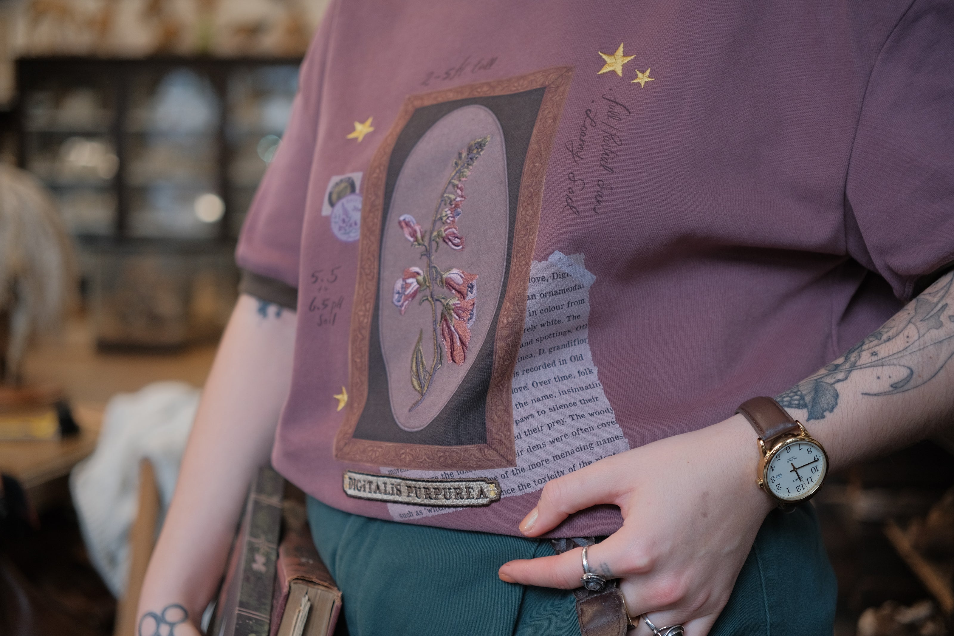Mauve-coloured cotton T-shirt featuring a detailed botanical illustration of a Digitalis Purpurea (foxglove) plant framed within an ornate oval design. The shirt is embellished with handwritten notes, stars, and vintage-style accents, creating a natural and artistic aesthetic.