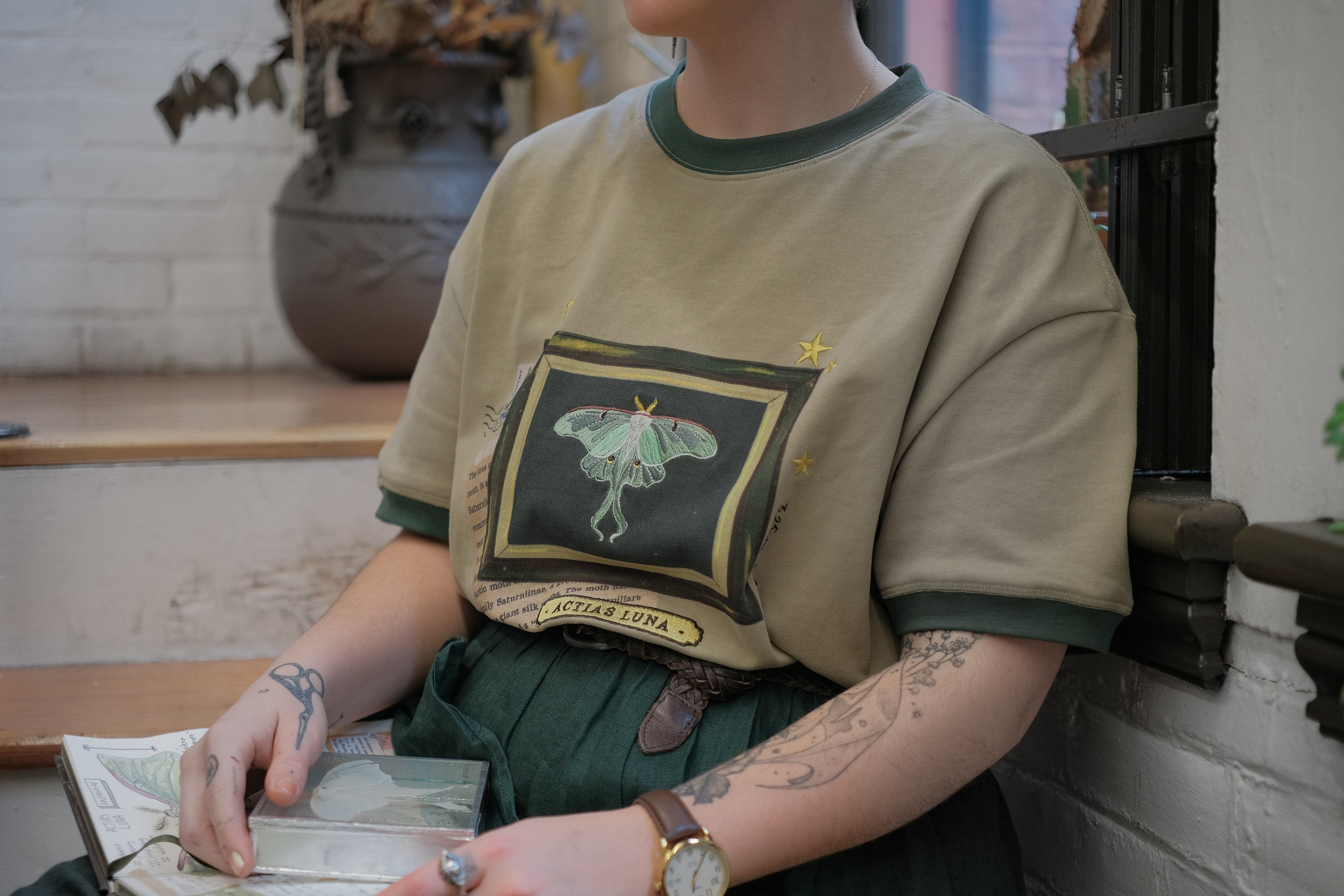 A light sea-foam green cotton T-shirt featuring an embroidered Luna moth design framed with vintage-style details, handwritten notes, celestial accents, and latin nomenclature.
