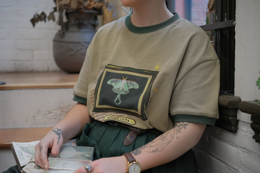 A light sea-foam green cotton T-shirt featuring an embroidered Luna moth design framed with vintage-style details, handwritten notes, celestial accents, and latin nomenclature.