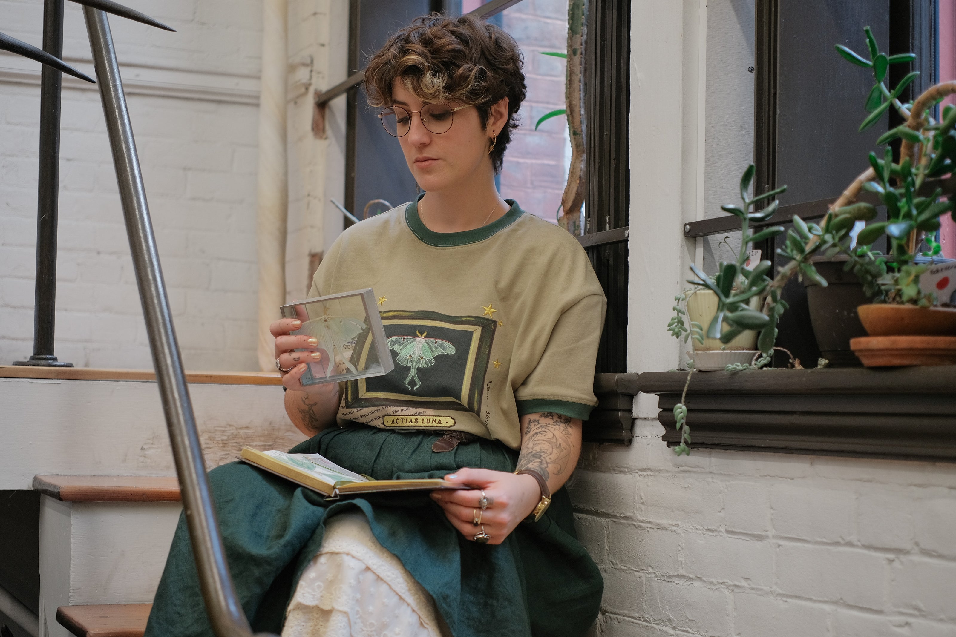A light sea-foam green cotton T-shirt featuring an embroidered Luna moth design framed with vintage-style details, handwritten notes, celestial accents, and latin nomenclature.