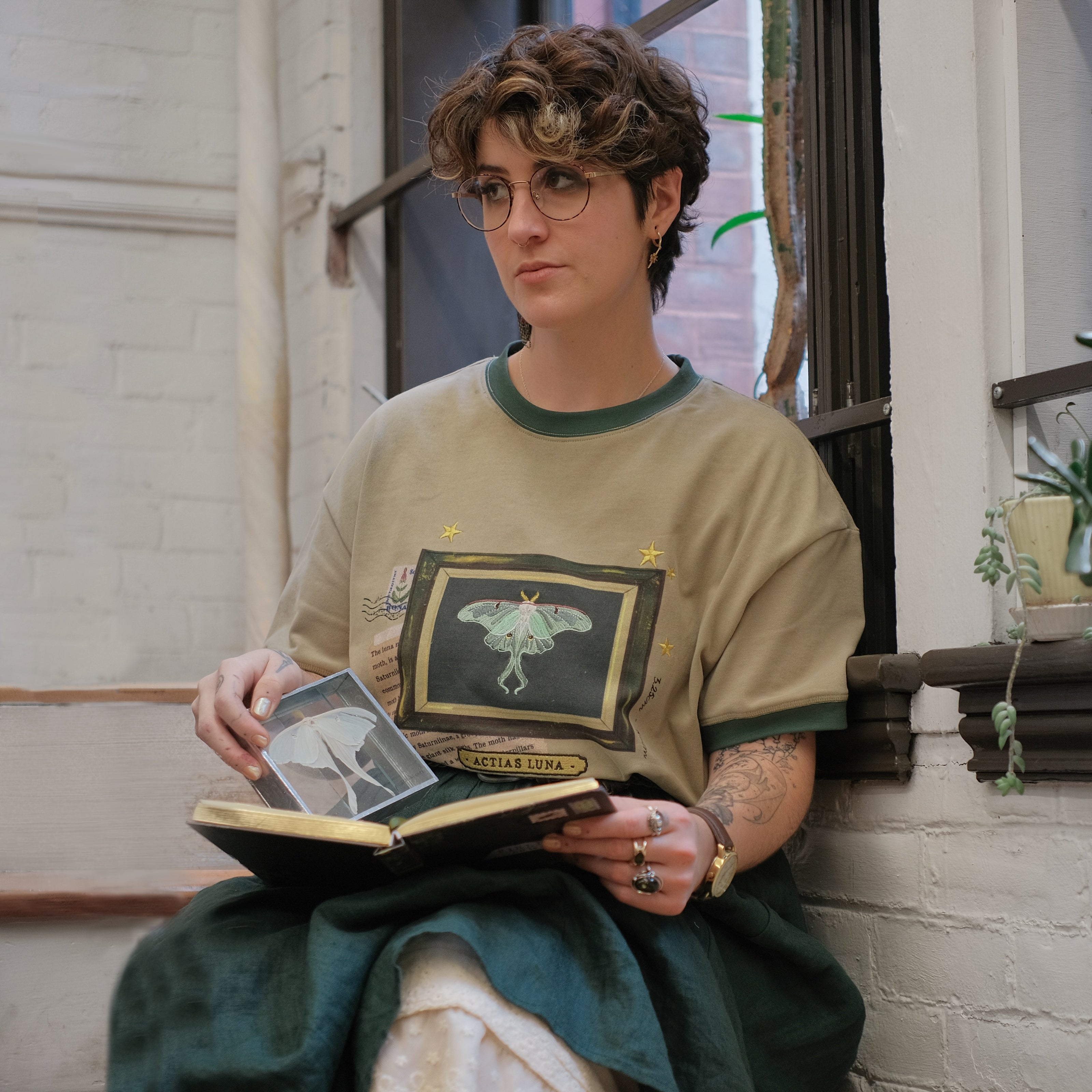 A light sea-foam green cotton T-shirt featuring an embroidered Luna moth design framed with vintage-style details, handwritten notes, celestial accents, and latin nomenclature.
