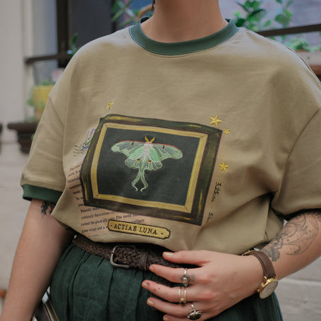 A light sea-foam green cotton T-shirt featuring an embroidered Luna moth design framed with vintage-style details, handwritten notes, celestial accents, and latin nomenclature.
