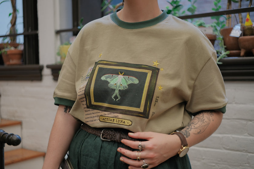 A light sea-foam green cotton T-shirt featuring an embroidered Luna moth design framed with vintage-style details, handwritten notes, celestial accents, and latin nomenclature.
