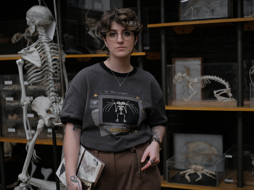 Charcoal gray cotton T-shirt featuring an embroidered skeletal illustration of Myotis lucifugus (a little brown bat) framed within a vintage-style display. The design includes handwritten text noting bat wingspan, golden star and moon accents, and a collage of vintage-inspired elements.