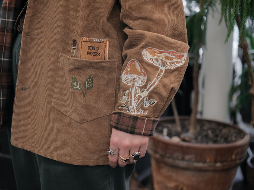 Light brown corduroy button up, Fall/spring jacket adorned with botanical and mycological illustrations, including mushrooms in a jar, latin nomenclature, and hand-drawn motifs. The jacket features green and red plaid accents on the collar and cuffs, along with functional pockets embroidered with nature-inspired designs.