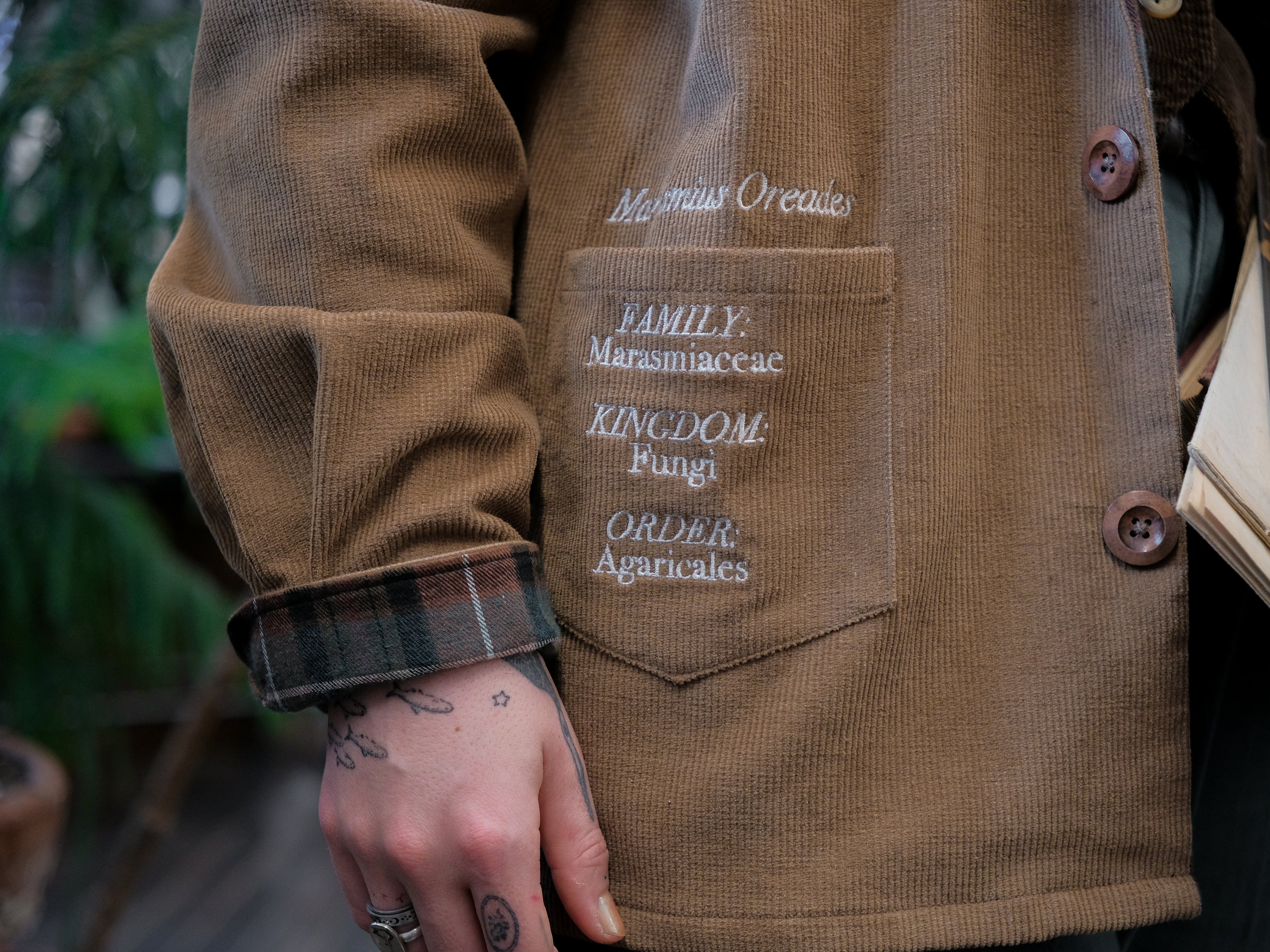 Light brown corduroy button up, Fall/spring jacket adorned with botanical and mycological illustrations, including mushrooms in a jar, latin nomenclature, and hand-drawn motifs. The jacket features green and red plaid accents on the collar and cuffs, along with functional pockets embroidered with nature-inspired designs.