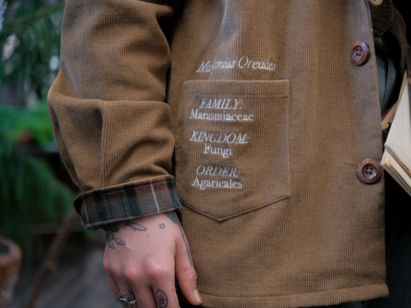 Light brown corduroy button up, Fall/spring jacket adorned with botanical and mycological illustrations, including mushrooms in a jar, latin nomenclature, and hand-drawn motifs. The jacket features green and red plaid accents on the collar and cuffs, along with functional pockets embroidered with nature-inspired designs.