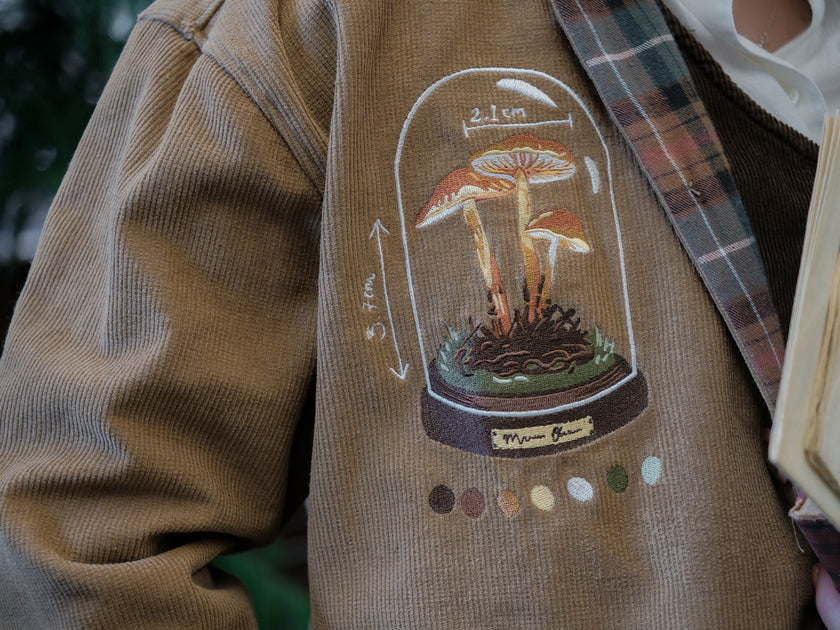 Light brown corduroy  button up, Fall/spring jacket adorned with botanical and mycological illustrations, including mushrooms in a jar, latin nomenclature, and hand-drawn motifs. The jacket features green and red plaid accents on the collar and cuffs, along with functional pockets embroidered with nature-inspired designs.