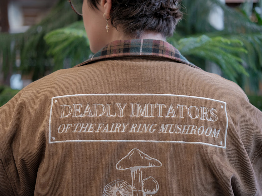 Light brown corduroy button up, Fall/spring jacket adorned with botanical and mycological illustrations, including mushrooms in a jar, latin nomenclature, and hand-drawn motifs. The jacket features green and red plaid accents on the collar and cuffs, along with functional pockets embroidered with nature-inspired designs.