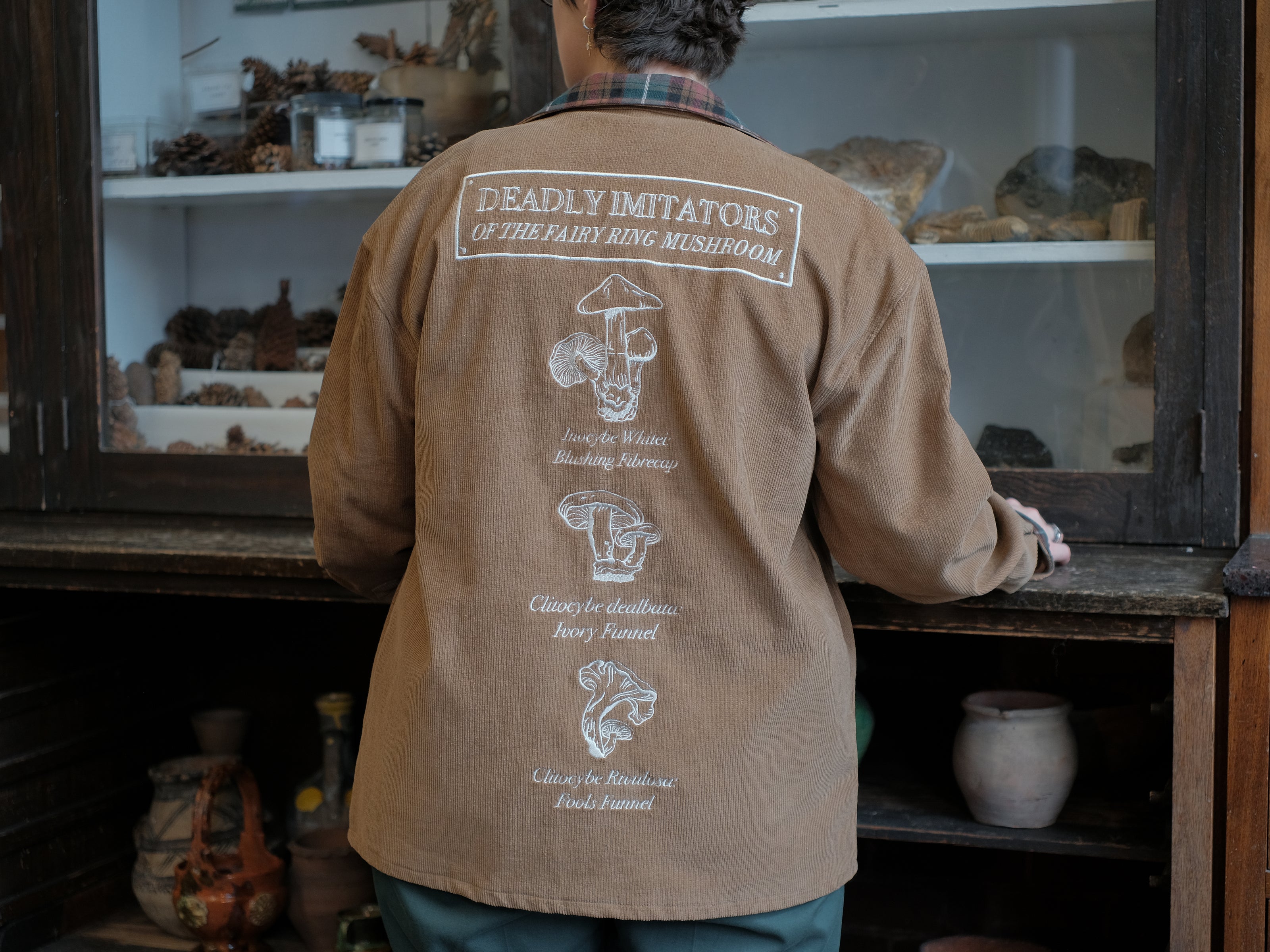 Light brown corduroy button up, Fall/spring jacket adorned with botanical and mycological illustrations, including mushrooms in a jar, latin nomenclature, and hand-drawn motifs. The jacket features green and red plaid accents on the collar and cuffs, along with functional pockets embroidered with nature-inspired designs.