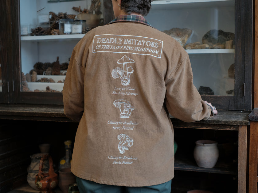 Light brown corduroy button up, Fall/spring jacket adorned with botanical and mycological illustrations, including mushrooms in a jar, latin nomenclature, and hand-drawn motifs. The jacket features green and red plaid accents on the collar and cuffs, along with functional pockets embroidered with nature-inspired designs.