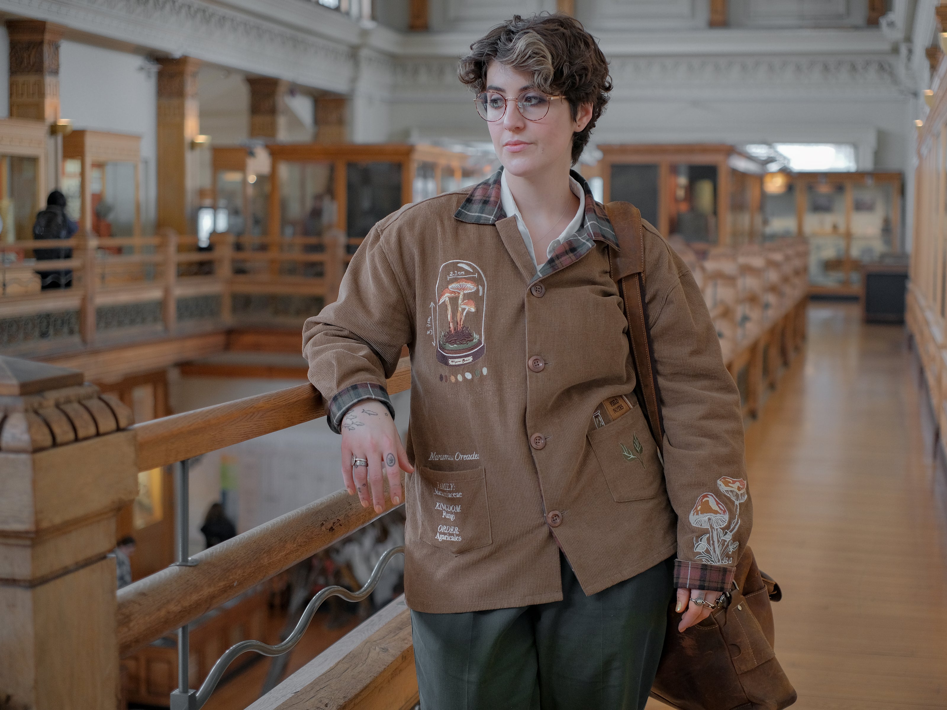 Light brown corduroy button up, Fall/spring jacket adorned with botanical and mycological illustrations, including mushrooms in a jar, latin nomenclature, and hand-drawn motifs. The jacket features green and red plaid accents on the collar and cuffs, along with functional pockets embroidered with nature-inspired designs.