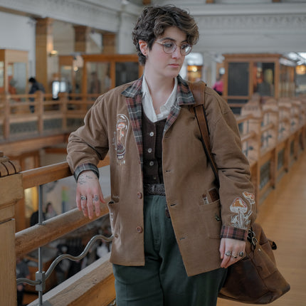Light brown corduroy button up, Fall/spring jacket adorned with botanical and mycological illustrations, including mushrooms in a jar, latin nomenclature, and hand-drawn motifs. The jacket features green and red plaid accents on the collar and cuffs, along with functional pockets embroidered with nature-inspired designs.