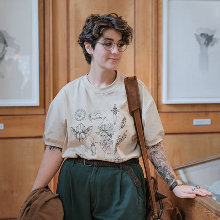 A light cream cotton T-shirt featuring black and gray vintage style sketches of specimens, including a rabbit foot, mandrake root, bat skull Luna moth, feather, stag-horn beetle, vial with a human tooth, star anise, thyme, and an orange slice.