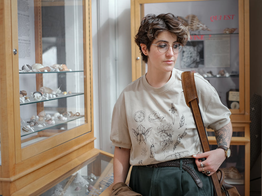 A light cream cotton T-shirt featuring black and gray vintage style sketches of specimens, including a rabbit foot, mandrake root, bat skull Luna moth, feather, stag-horn beetle, vial with a human tooth, star anise, thyme, and an orange slice.