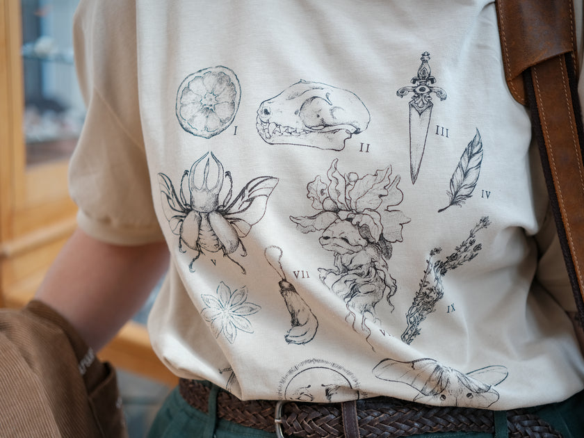 A light cream cotton T-shirt featuring black and gray vintage style sketches of specimens, including a rabbit foot, mandrake root, bat skull Luna moth, feather, stag-horn beetle, vial with a human tooth, star anise, thyme, and an orange slice.