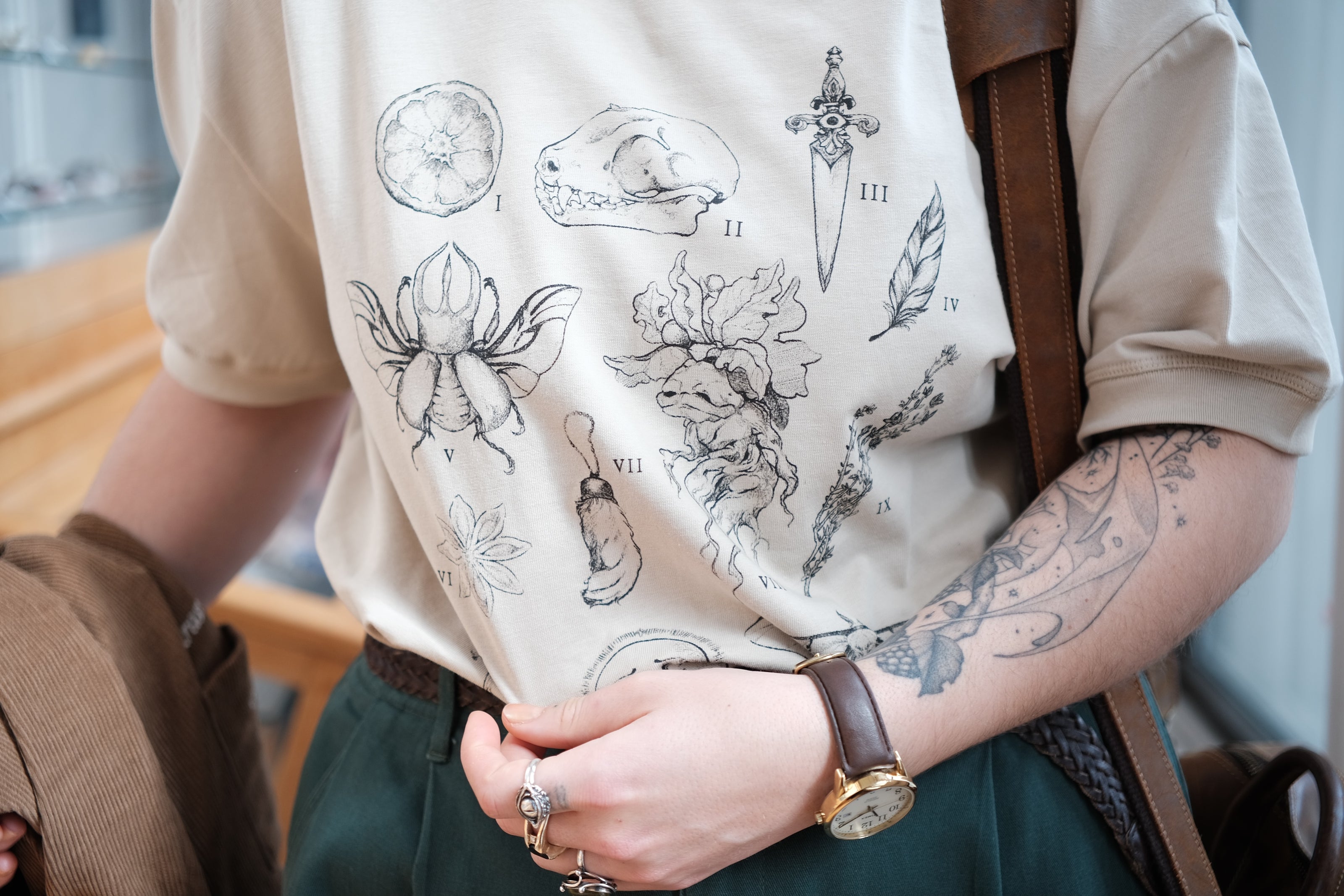 A light cream cotton T-shirt featuring black and gray vintage style sketches of specimens, including a rabbit foot, mandrake root, bat skull Luna moth, feather, stag-horn beetle, vial with a human tooth, star anise, thyme, and an orange slice.