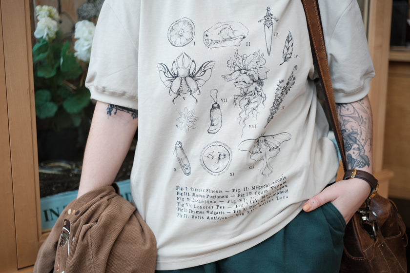 A light cream cotton T-shirt featuring black and gray vintage style sketches of specimens, including a rabbit foot, mandrake root, bat skull Luna moth, feather, stag-horn beetle, vial with a human tooth, star anise, thyme, and an orange slice.