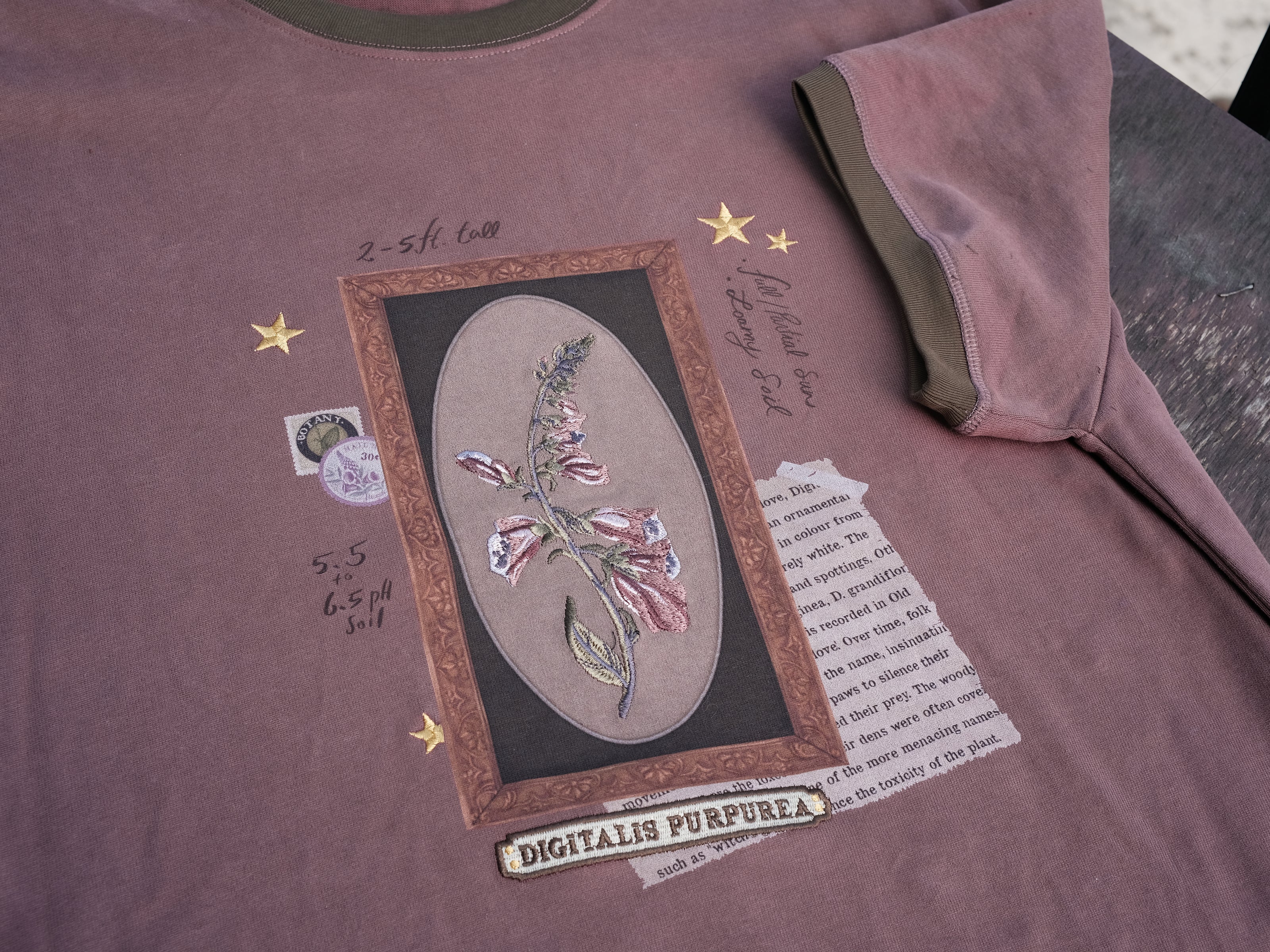 Mauve-coloured cotton T-shirt featuring a detailed botanical illustration of a Digitalis Purpurea (foxglove) plant framed within an ornate oval design. The shirt is embellished with handwritten notes, stars, and vintage-style accents, creating a natural and artistic aesthetic.