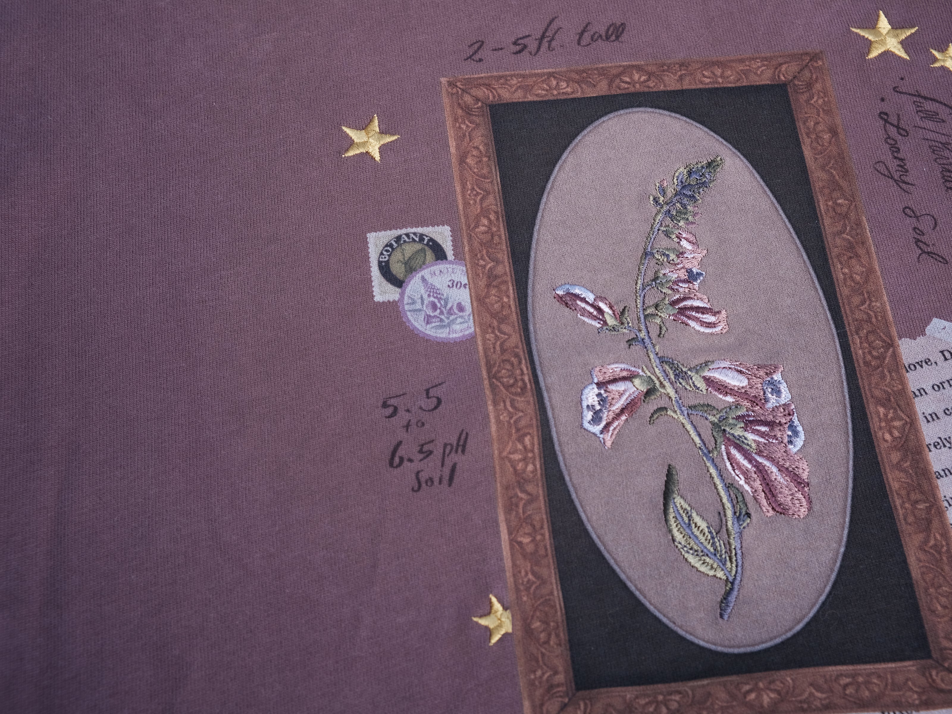 Mauve-coloured cotton T-shirt featuring a detailed botanical illustration of a Digitalis Purpurea (foxglove) plant framed within an ornate oval design. The shirt is embellished with handwritten notes, stars, and vintage-style accents, creating a natural and artistic aesthetic.