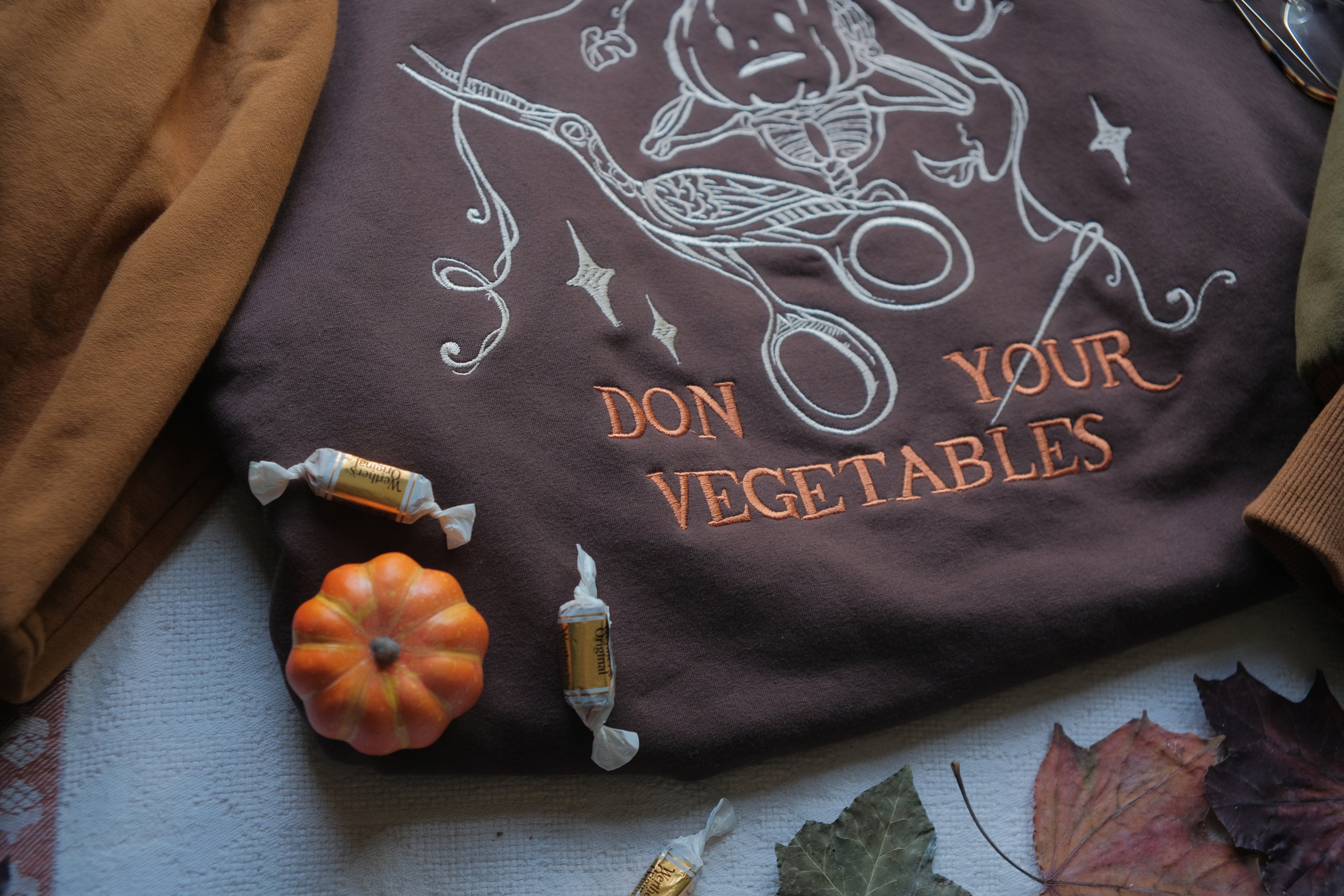 Cozy, oversized, puff-sleeved, cotton crewneck sweater in pumpkin-orange and shades of brown. The sweater features an intricately embroidered design in white and orange, of a pumpkin skeleton, vines, stork scissors, and reads â€œDon Your Vegetablesâ€.