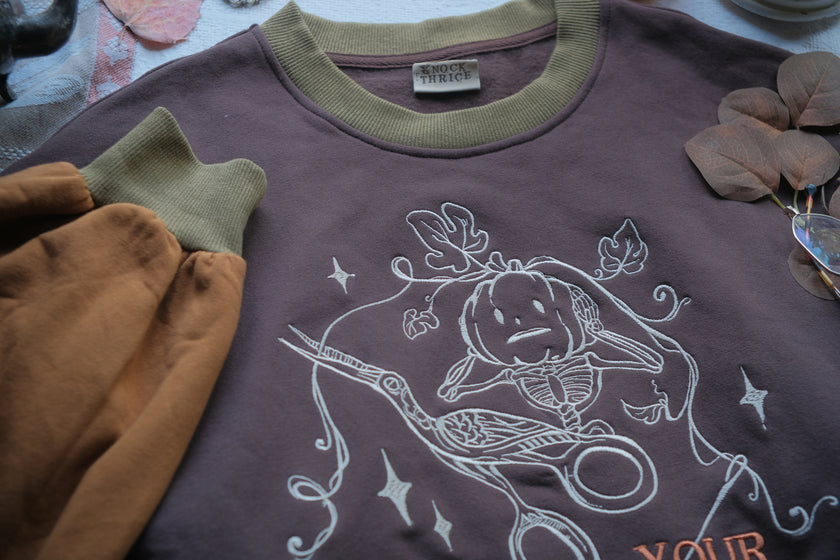 Cozy, oversized, puff-sleeved, cotton crewneck sweater in pumpkin-orange and shades of brown. The sweater features an intricately embroidered design in white and orange, of a pumpkin skeleton, vines, stork scissors, and reads â€œDon Your Vegetablesâ€.