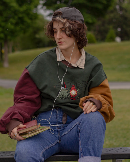 Cozy, oversized, puff-sleeved, color-blocked, cotton crewneck sweater in forest green, with burgundy, and mustard coloured sleeves. The sweater features an intricately embroidered design of a green frog with a red wizard hat and a red lantern on the chest.