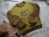 Cozy, oversized, puff-sleeved, color-blocked, cotton crewneck sweater in swamp-green, and light and dark brown. The sweater features an intricately embroidered design of a green frog with a red wizard hat and a red lantern on the chest.