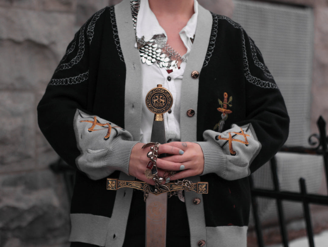 A soft button-up black and silver knit cardigan. With black and gold moon buttons, the design features intricate embroidery of a vine wrapped sword, chainmail, and armour. Evoking a whimsical and fantasy-inspired look.