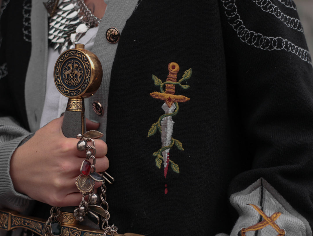 A soft button-up black and silver knit cardigan. With black and gold moon buttons, the design features intricate embroidery of a vine wrapped sword, chainmail, and armour. Evoking a whimsical and fantasy-inspired look.