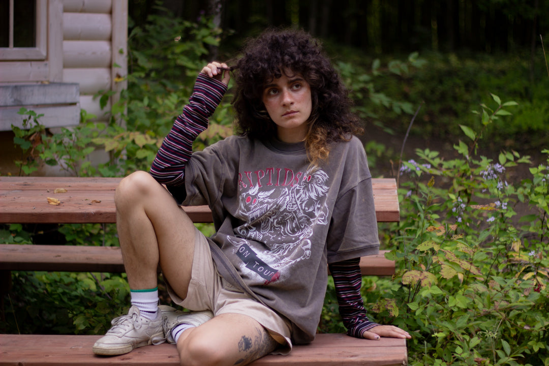 Bone-dust brown oversized cotton 80â€™s style band t-shirt with a vintage-style graphic depicting cryptids, including Bigfoot and Mothman, along with the bold text 'Cryptids On Tour' in red and white, and a list of shows across various locations and dates on the back.