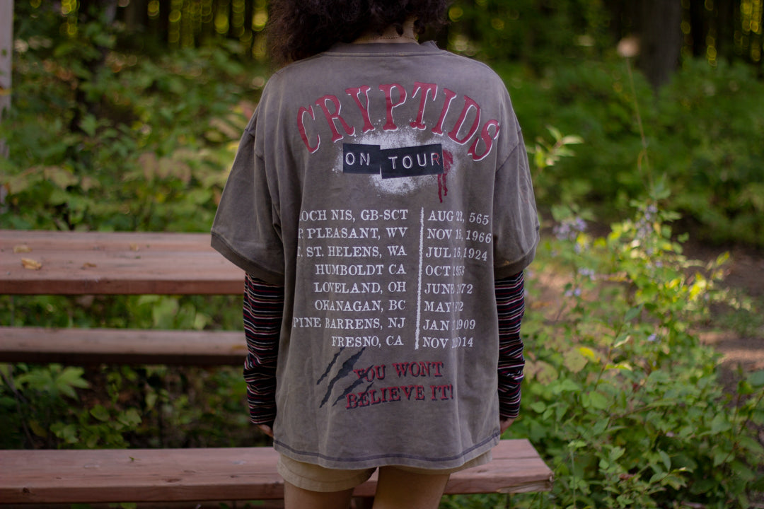 Bone-dust brown oversized cotton 80â€™s style band t-shirt with a vintage-style graphic depicting cryptids, including Bigfoot and Mothman, along with the bold text 'Cryptids On Tour' in red and white, and a list of shows across various locations and dates on the back.