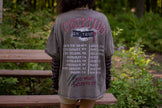 Bone-dust brown oversized cotton 80’s style band t-shirt with a vintage-style graphic depicting cryptids, including Bigfoot and Mothman, along with the bold text 'Cryptids On Tour' in red and white, and a list of shows across various locations and dates on the back.