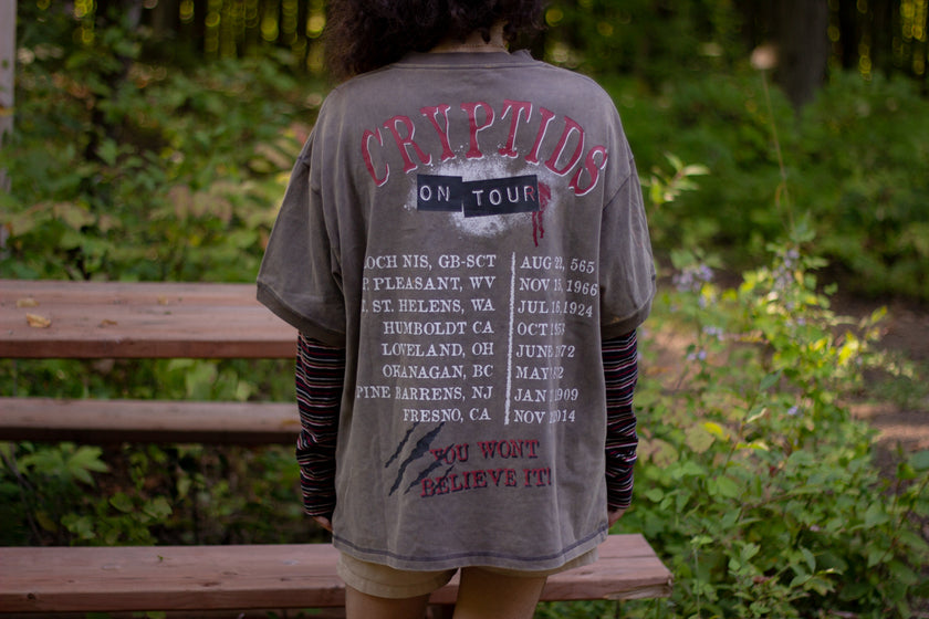 Bone-dust brown oversized cotton 80â€™s style band t-shirt with a vintage-style graphic depicting cryptids, including Bigfoot and Mothman, along with the bold text 'Cryptids On Tour' in red and white, and a list of shows across various locations and dates on the back.