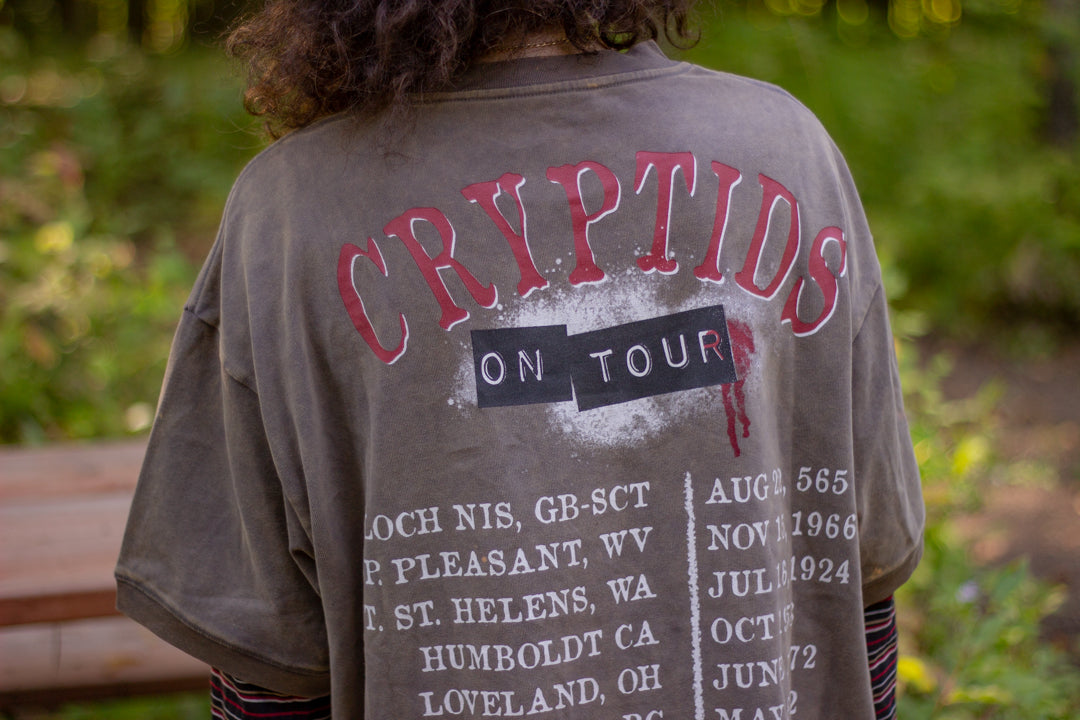 Bone-dust brown oversized cotton 80’s style band t-shirt with a vintage-style graphic depicting cryptids, including Bigfoot and Mothman, along with the bold text 'Cryptids On Tour' in red and white, and a list of shows across various locations and dates on the back.