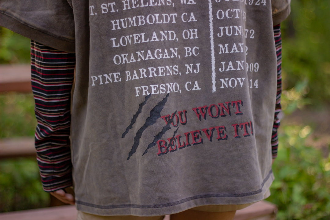 Bone-dust brown oversized cotton 80â€™s style band t-shirt with a vintage-style graphic depicting cryptids, including Bigfoot and Mothman, along with the bold text 'Cryptids On Tour' in red and white, and a list of shows across various locations and dates on the back.