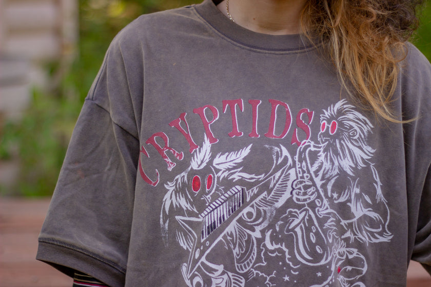 Bone-dust brown oversized cotton 80â€™s style band t-shirt with a vintage-style graphic depicting cryptids, including Bigfoot and Mothman, along with the bold text 'Cryptids On Tour' in red and white, and a list of shows across various locations and dates on the back.