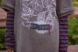 Bone-dust brown oversized cotton 80’s style band t-shirt with a vintage-style graphic depicting cryptids, including Bigfoot and Mothman, along with the bold text 'Cryptids On Tour' in red and white, and a list of shows across various locations and dates on the back.