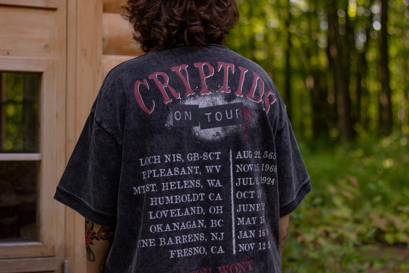 Black acid-washed oversized 80's style cotton band t-shirt with a vintage-style graphic depicting cryptids, including Bigfoot and Mothman, along with the bold text 'Cryptids On Tour' in red and white, and a list of shows across various locations and dates on the back.